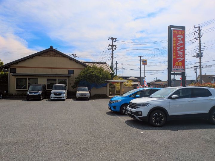 プレミアラーメンショップ久松山倉坂下2号店の店外にある赤い看板と、駐車場の様子。