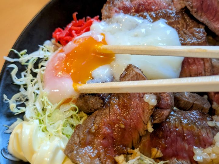 ステーキ丼に乗せた温泉卵の黄身を箸で割って、お肉に絡めている様子。